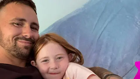 Family handout Nyla and her dad together on a sofa. Kieran is dressed in dark clothing and has a tattooed arm wrapped gently around Nyla. Nyla is leaning in against him, wearing a light‑coloured T‑shirt with a small design on the front and dark trousers. They are seated on a soft, blue‑covered sofa, and a bright pink object is visible to the right side of the frame.