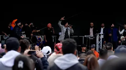 A man in a turban stands on a stage with a microphone in one hand and his other hand in the air. Around 10 other men stand on stage with various instruments or microphones. They stand in front of a crowd, who are watching the stage.