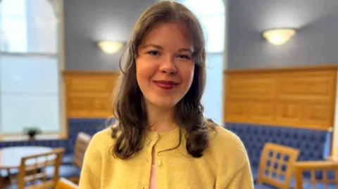 Freya has medium length brown wavu hair worn down. She is wearing a yellow cardigan. She is standing in a room panelled in pine wood with blue at the top part of the wall. At the bottom is banquette seating in blue leather and several chairs behind her.
