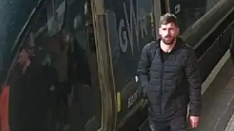 CCTV image of Lee Horton next to a GWR train. He is standing on the platform next to the train. He is wearing a black jacket. He has a beard and short hair.