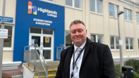 Rob looks at the camera as he stands outside Highlands College. He is wearing a long smart black coat. He has a dotted shirt on with a patterned tie. Rob has short grey hair.