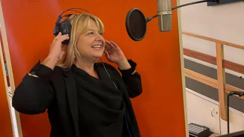 Kerr Family A blonde woman stands in a recording studio wearing headphones, positioned in front of a suspended microphone with a pop filter. Bright lighting and orange acoustic panels surround the setup, with a music stand visible in the foreground. The woman is smiling as she speaks or sings.