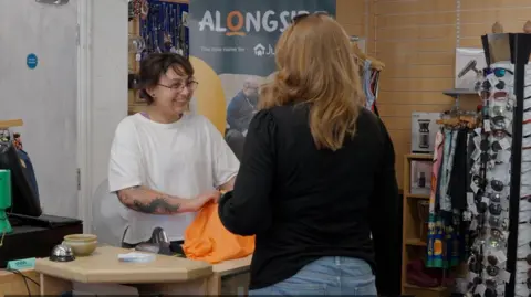A woman with tattoos on her arms wearing a short-sleeved white t-shirt is serving a customer in a charity shop. On the right is a stand with sunglasses. 