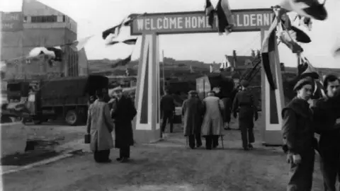 A black and white historical photograph. It shows bunting and an arch that reads "welcome home". An elderly couple with walking sticks are seen passing through the arch. There are others standing around and a military truck is visible.