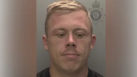A police custody photo of McCaulay Parker who has short blond hair and is wearing a black t-shirt. He is looking down. The wall behind him is grey.