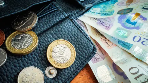 A stock image of a purse with Sterling coins coming out of it and several bank notes. 