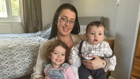 Chanel with her two children. One is a toddler and the other is five months old. They are all smiling at the camera. There is a bed behind them.