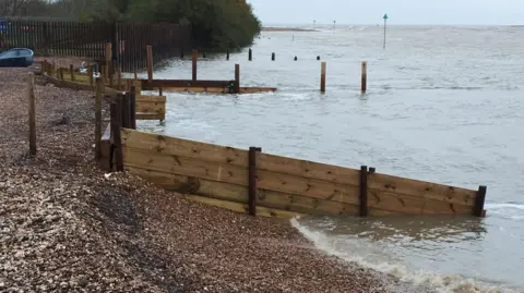 Bawdsey Haven Yacht Club An image of the coastline, where the beach meets the sea. Wooden groynes and trees in the distance also feature in the image.