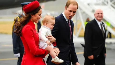 The Princess of Wales, wearing a red coat, smiles as she carries baby Prince George, dressed in white and wearing cream baby shoes.