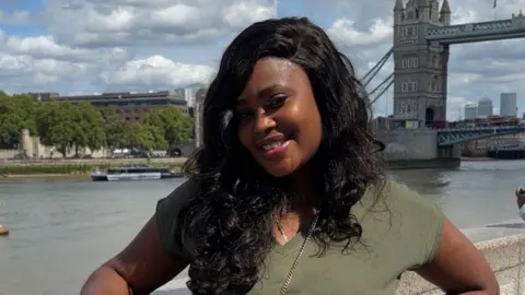 Georgina Tuffour A head-and-shoulders shot of Georgina Tuffour standing by the River Thames in front of Tower Bridge. She is wearing a green t-shirt with a handbag over one shoulder.