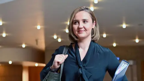 Getty Images Mairi McAllan - a young woman with shoulder-length blonde hair, wearing a dark blouse and carrying a bag over her shoulder and a blue folder under her arm, pictured in the Scottish Parliament
