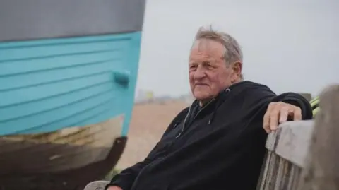 A man wearing a black hooded top and grey shorts. He is sat on a bench with a blue boat on a pebbled beach behind him.