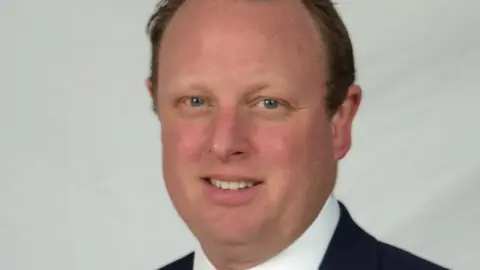 Borough Council of King's Lynn and West Norfolk A man looks at the camera in a formal photograph. He has thinning brown hair, cut short, and blue eyes. 