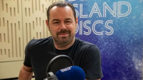 Danny Dyer sits smiling in front of a Desert Island Discs backdrop.