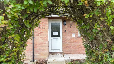 Ben Schofield/BBC The front door of a house on Clifden Close. The door is white, UPVc, with a glass pane, metal handle and taped-up letter box. A hand-written signed taped to the glass says "wet latex". An outside light is mounted to the right of the door, as an empty hanging basket bracket. The image is framed by the branches and leaves of two bushes that have been trained to grow into an arch shape. A concrete footpath leads from the bottom middle of the picture to the front door. Another path leads from the door and heads off to the right. The red bricks of the front of the house surround the door.
