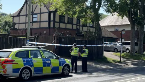 A police car and two police officers are stood outside crime tape which is in front of a large gate, in a residential area. 
