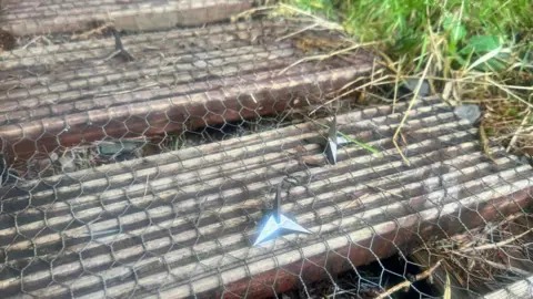 North Argyll Cycle Club Two metal throwing stars resting on chicken wire laid over stacked, weathered wooden logs outdoors, with grass visible at the edge