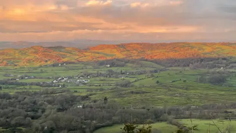 BBC Weather Watchers/Gee A valley of fields with trees and clusters of houses. The hills in the background are cast in an orange light. The sky is covered in white and grey clouds.