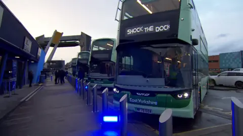 Several green buses are lined up outside The Deep aquarium, one of them is displaying 'shock the dock' on its display board.