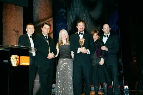 Geoff Thompson Four men and two women stand on stage next to a podium which has a golden BAFTA face attached to it alongside a logo for the phone company orange. The four men wear black tuxedos. A blonde woman in the middle wears a black top and long grey floral skirt. A woman on the right wears a black top and skirt.