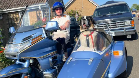 John Devine/BBC Alan Valkeith is sitting on a blue motorbike which has a sidecar with a St Bernard dog, called Louis, sitting in it. Valkeith is wearing a white T-shirt and a blue helmet, and there is a bungalow and van in the background.