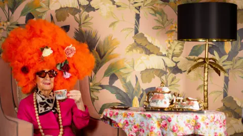 Maplewood Court Care Home The image shows an elderly woman sat in a chair, wearing a bright orange wig with roses in them. She is wearing a large pearl necklace and holding a teacup. The wallpaper in the background is covered in leaves and a black and gold lamp stands on a table to the right, along with an ornate tea pot and cups.