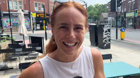 BBC/NATHAN TURVEY A picture of local resident Amy Young who has ginger hair, is wearing a white top and is smiling - there are shops and a road in the background.
