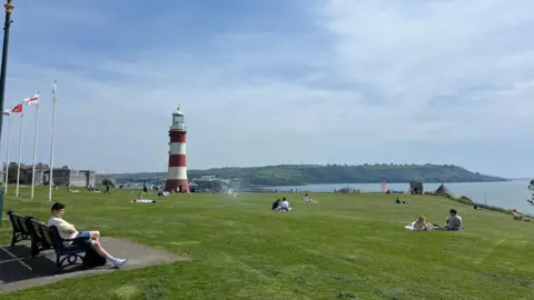 A general view of Plymouth Hoe. There is a large grass area, with a lighthouse in the distance. People are sat in groups on the grass and in the distance is the sea and land.