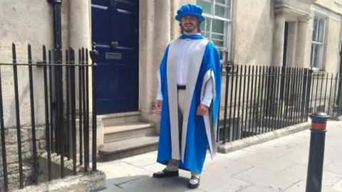 Eddie Ilic A man wearing blue and white doctorate robes, and a blue cap. He is photographed on a street in Bath smiling