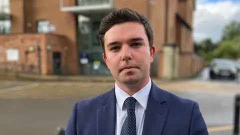 A man with short dark hair looks at the camera. He is wearing a white shirt with a polka dot navy and white tie and a navy blue blazer. There is a brown bricked building behind him, as he is standing in a car park. You can see blurred in the background yellow lines on the ground for parking spaces.