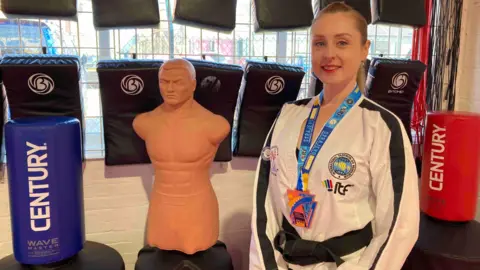 A woman wearing a white jacket, trousers and a black belt, along with a lanyard, stands in a martial arts studio. Various equipment, including punching bags, cushions and a dummy can be seen behind her.
