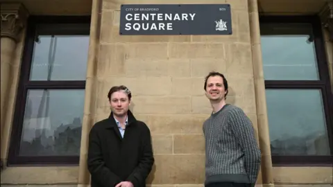 Two men stand below a smart black sign saying City of Bradford Centenary Square, which also features Bradford's civic crest