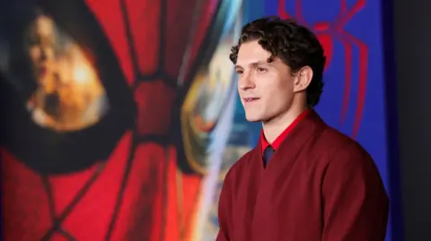 PA Media A man wearing a red jumper looks past the camera with a neutral look. Behind him is a giant Spider-Man mask looming over him.