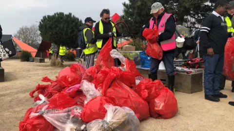 A picture of a small pile of red and clear bin bags filled with waste. They are gathered together on the sand in Cardiff Bay. Five men stand behind the bin bags wearing high vis vests in fluorescent- yellow and pink. Trees can be seen in the distance.  