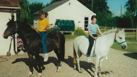 March family A boy aged about 11 in a yellow T-shirt and blue jeans looks down while sitting on a brown horse, while beside him a girl aged about nine in a pale blue T-shirt and black trousers smiles at the camera while sitting on a white horse.