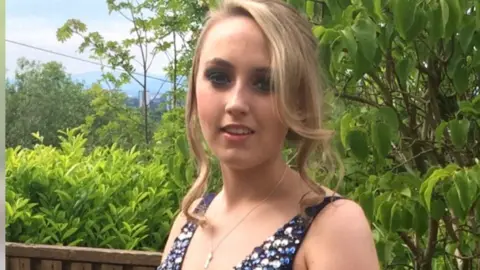 Katie Allan has long blonde hair tied back. She smiles at the camera and stands in front of green leafy trees and wears a silver and blue bejewelled top.