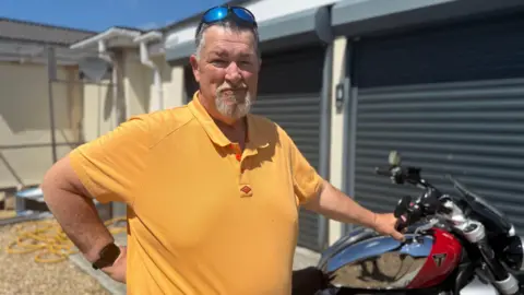 BBC A man with short dark grey hair and a white goatee beard looking at the camera. He is wearing a mustard yellow polo and has sunglasses resting on his head. You can see a red and silver motorbike in the background
