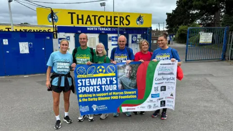 Darby Rimmer Foundation Six people holding a banner in front of a football stadium. The banner read 'Stewart's Sports Tour'