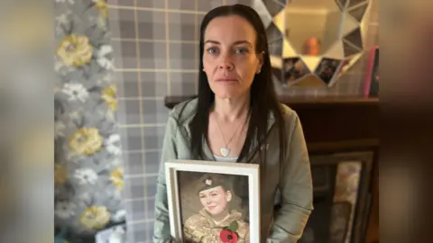 Family A woman with long black hair is standing in a living room holding a framed picture of a teenage girl dressed in an Army cadet uniform. The girl is wearing Army fatigues and a beret. The woman is wearing a light-green sports top and has a neutral expression on her face. In the background is a mirror over a fireplace and grey and yellow patterned wallpaper.