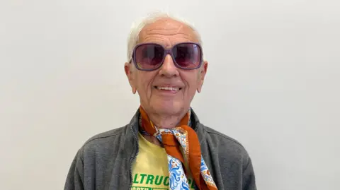 Bob Emmerson wearing dark eyeglasses and standing against a white wall looks directly at the camera.