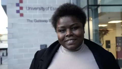 John Fairhall/BBC Ojoma Idegwu smiles as she looks to the camera in front of the University of Essex Southend campus. She has short black hair and wears a black coat with a black jumper underneath. 