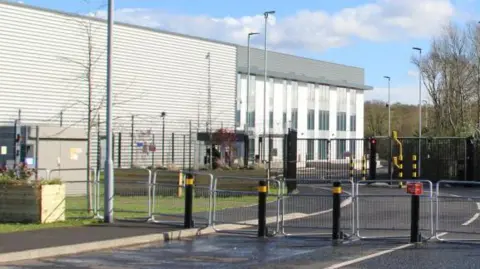 The exterior of an industrial building. There are bollards and fencing across the road, and a large warehouse behind large gates. 