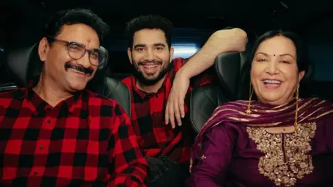 Samay Raina via Instagram Samay Raina poses gleefully with his mum and dad inside a car. He and his father are wearing matching red-black check shirts, while his mum is wearing a purple tunic and scarf with golden threadwork. 