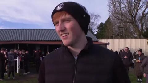 Angry Ginge wears a beanie woolly hat covering his ginger hair. He is smiling and wearing warm clothing. People are behind him at the football club, also wrapped up warm.
