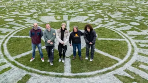 Walk the Plank Four smiling people are standing in the central circle of the artwork, made using white marker, that contains the words "Heart of Ashington". It depicts lots of symbols including of ash branches, footballs and miners' wheels.