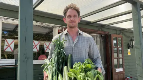A man standing under a covered porch holds a large wicker basket containing cucumbers, spring onions and leafy green vegetables outside a green-painted farm shop building.