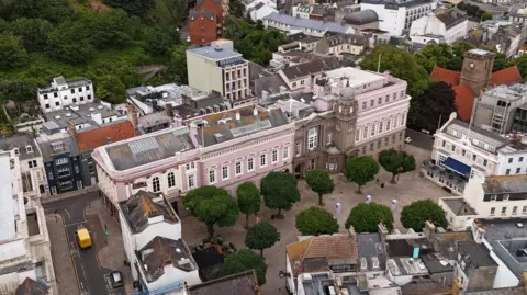 BBC An aerial image of a long building made from pink granite, with white windows. A number of green trees are visible in front of the building.