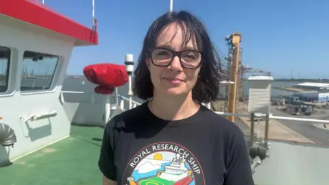 Richard Knights/BBC Dr Hogan wears a black T-shirt with a picture of the RSS Sir David Attenborough. She is standing on the deck of the ship. 