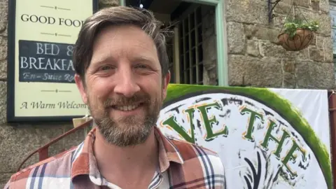 Sam Fitch is smiling at the camera as he stands in front of the pub's entrance with a welcome sign and a Save our Stag sign behind him. He has brown short hair and a beard and moustache and is wearing an orange, white and black checked shirt.