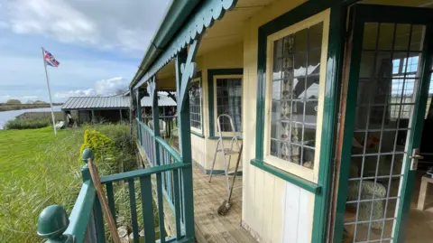 Shaun Whitmore/BBC The view from Towerview's veranda, which is painted green wood. The walls of the house are cream painted wood and it has a wooden floor. Beyond the house is grass, with a flagpole flying a Union Flag, and beyond that can be seen the Broads waterway